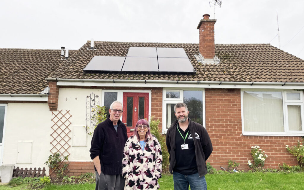 Two men and a woman standing outside red brick bungalow with solar panels on roof.