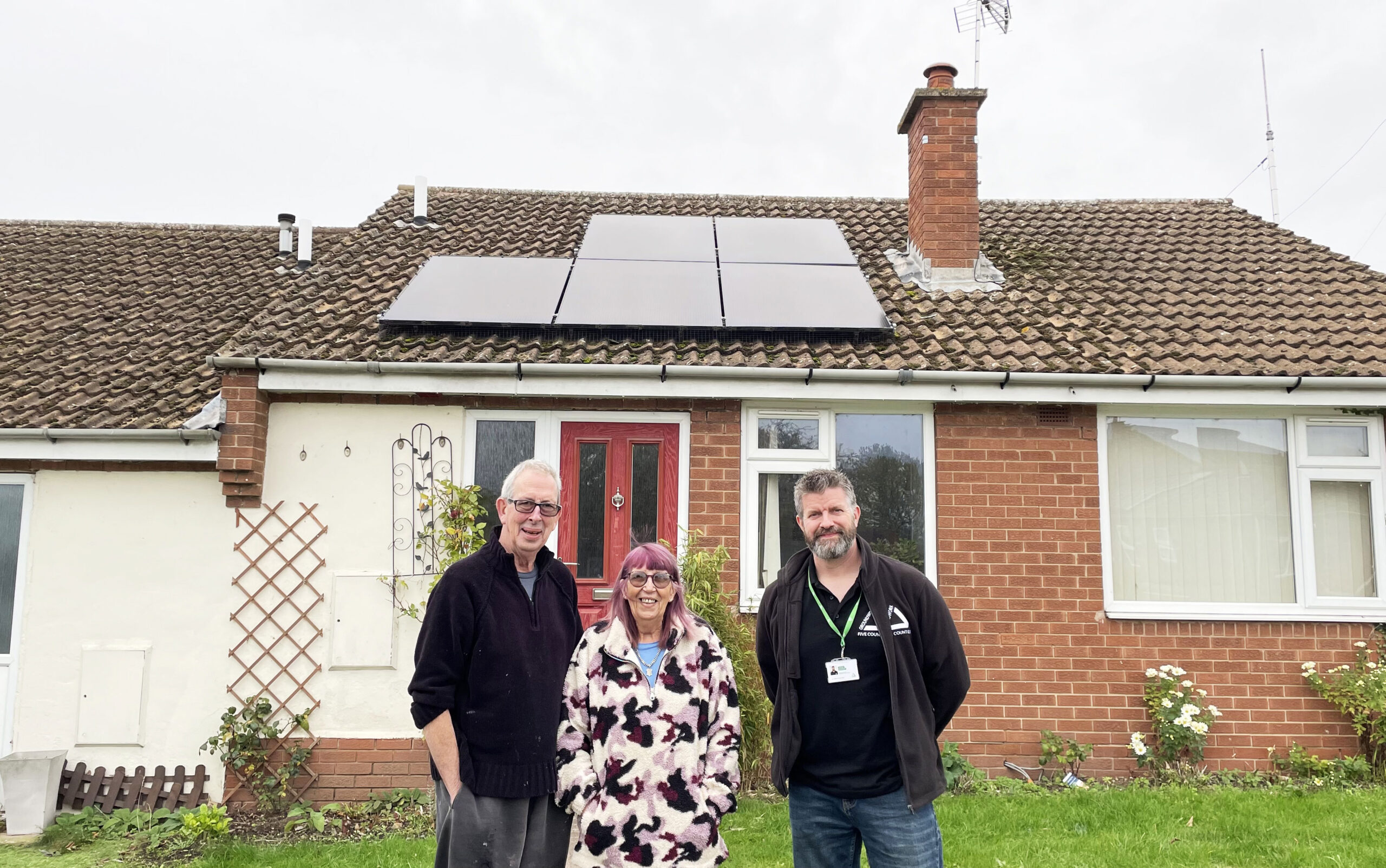Two men and a woman standing outside red brick bungalow with solar panels on roof.