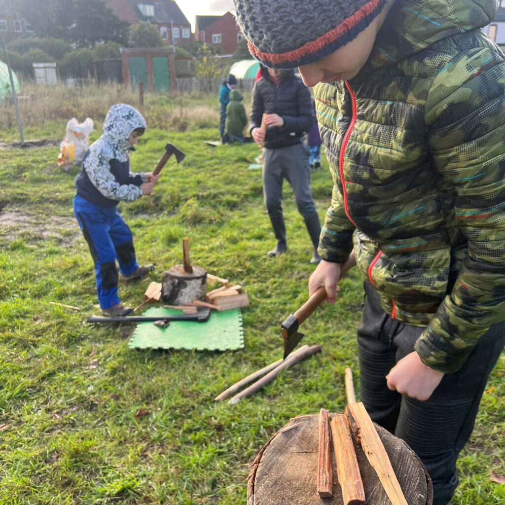 Young man with small axe and bits of wood, in the background are people chopping wood in outdoor setting