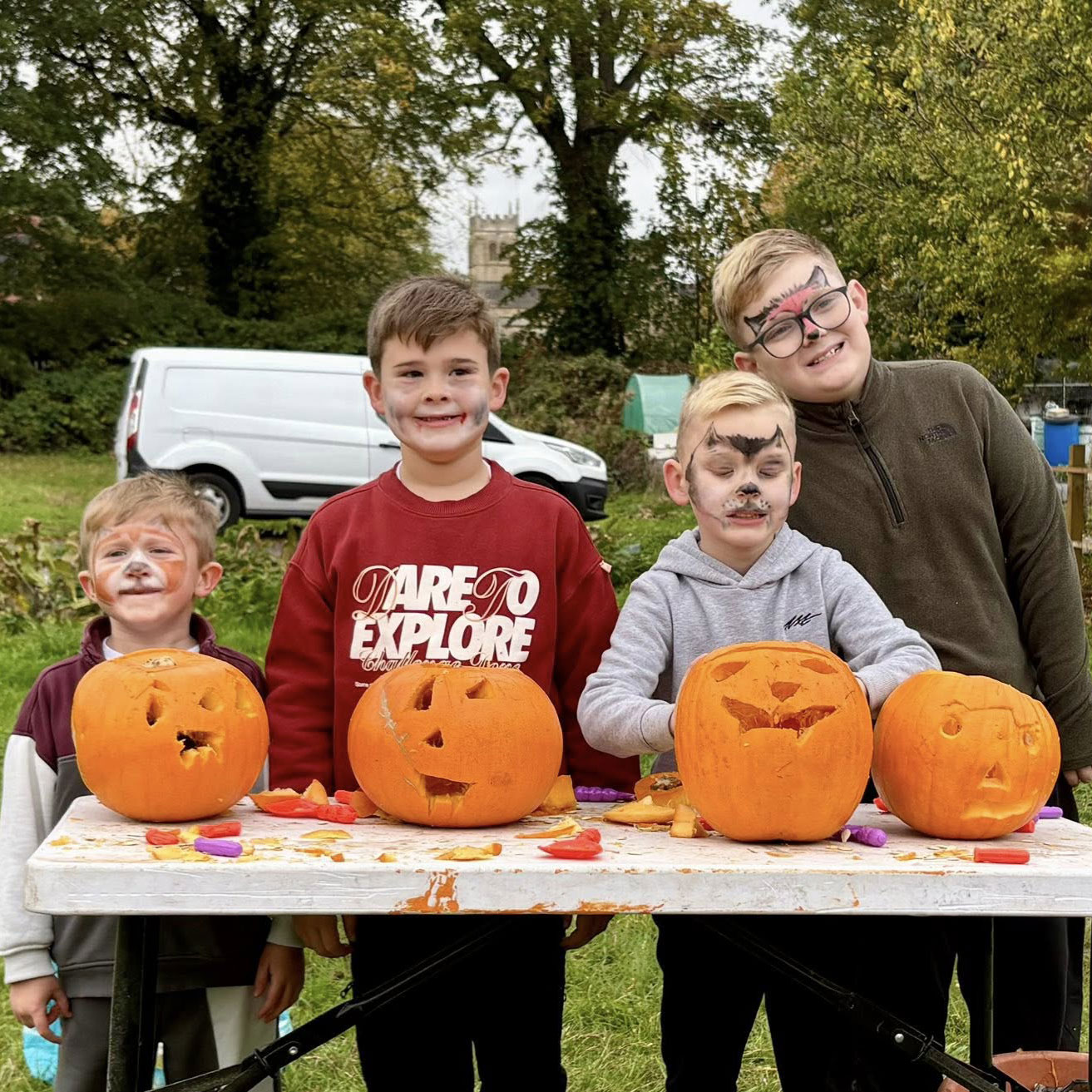 Four young boys with face paint with carved pumpkins on white table in outdoor setting.