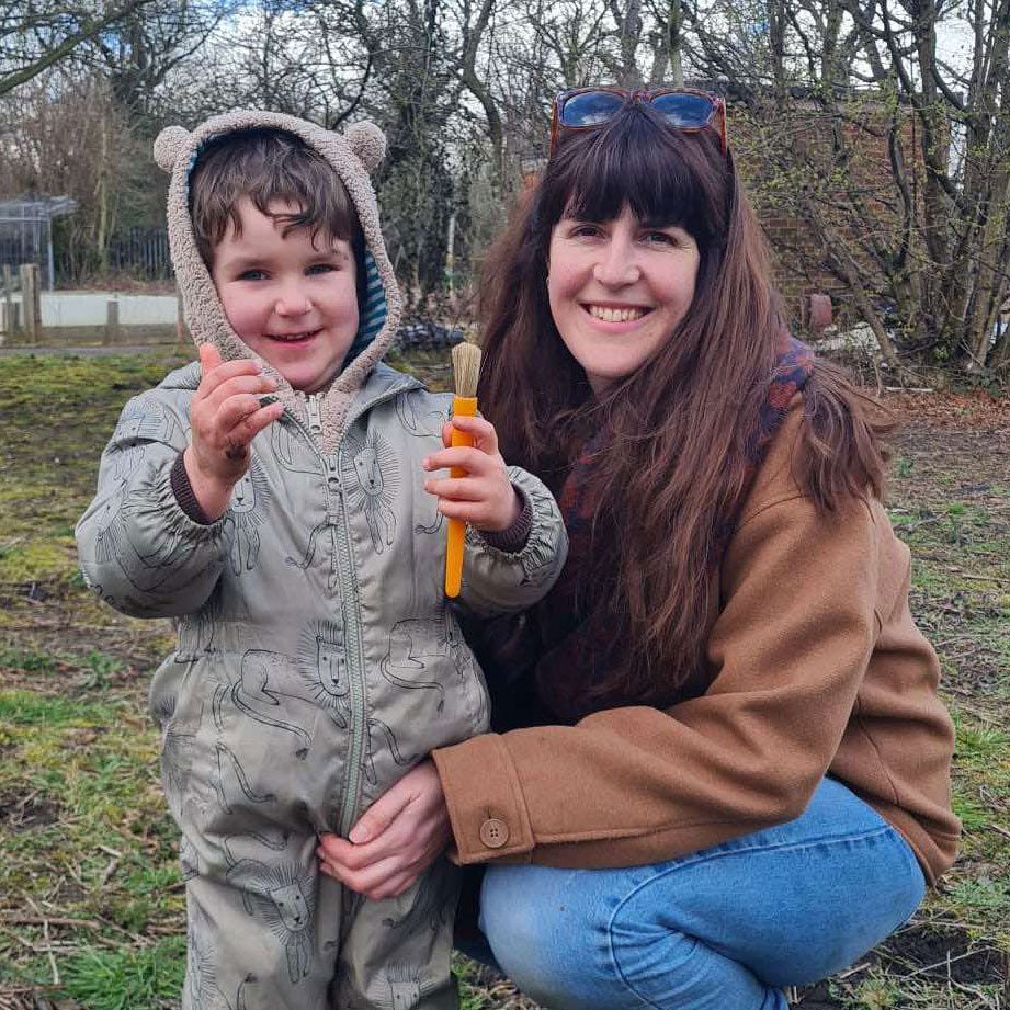 Brunette woman and young boy wearing grey onesie holding brush in outdoor setting and smiling.