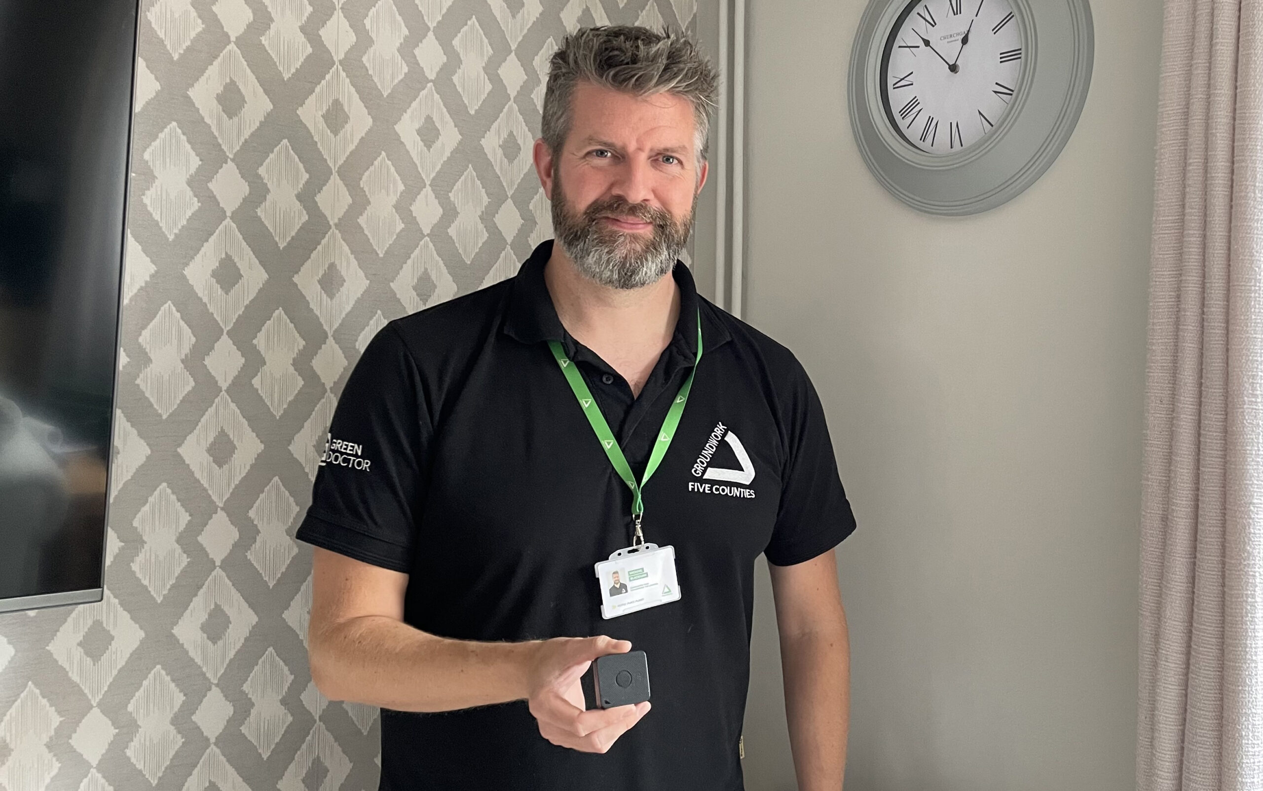 Man wearing black polo shirt and green lanyard holding small black rectangular item, standing by wallpapered wall and clock