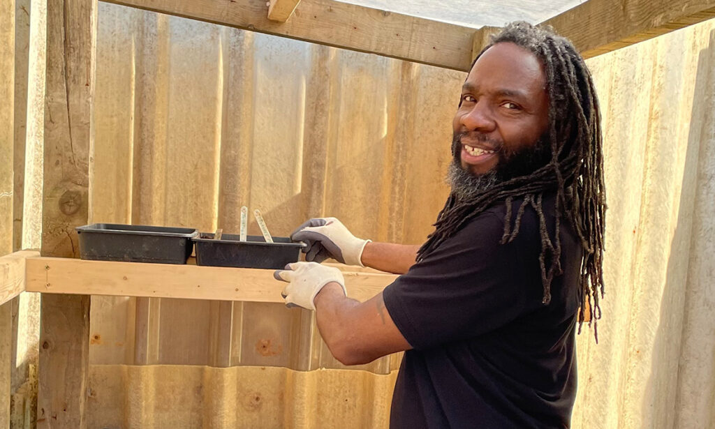 Black man with locs wearing white gloves and dark T-shirt in greenhouse with two plastic tubs.