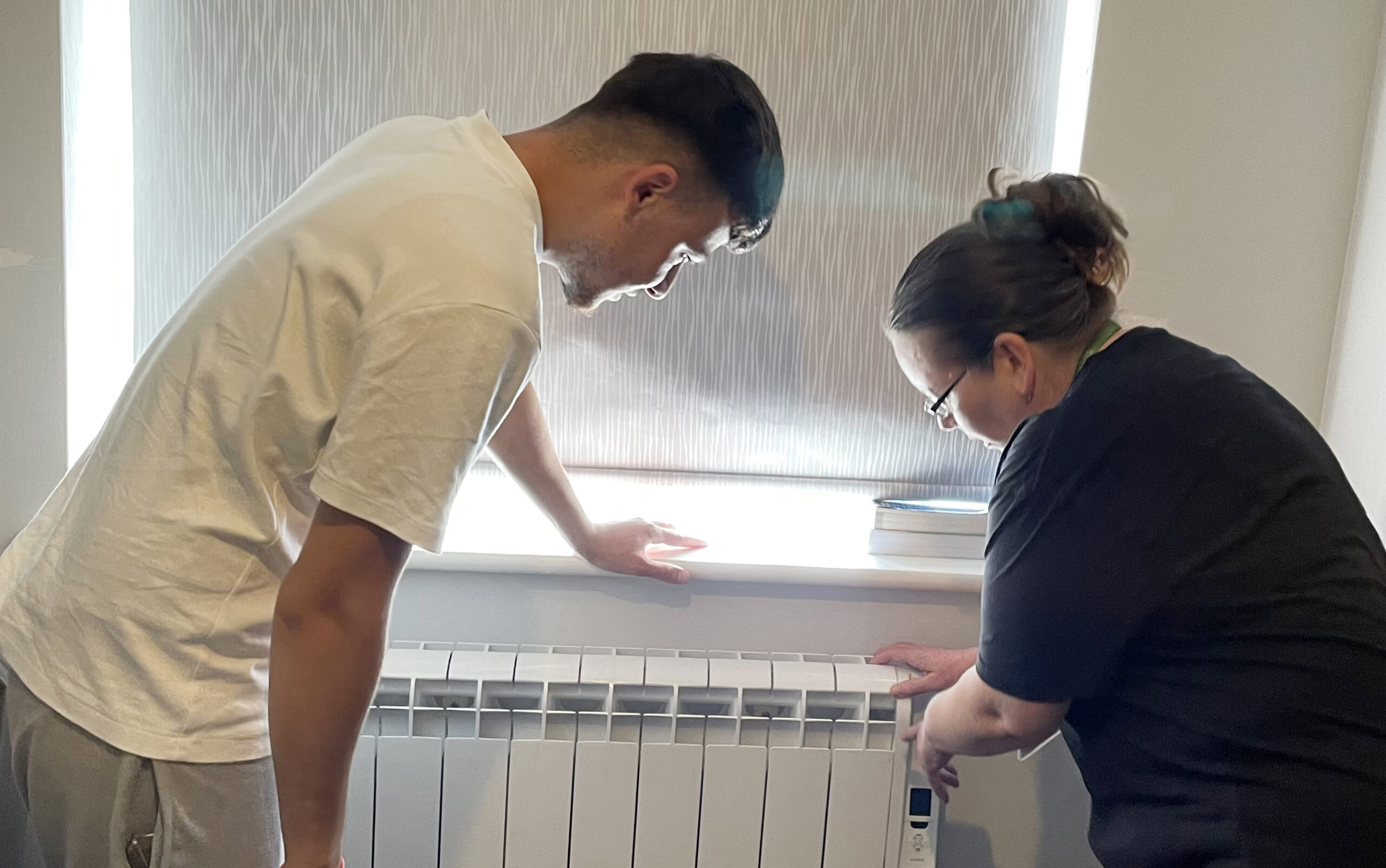 Man wearing white T-shirt and woman wearing black looking at a radiator underneath window.