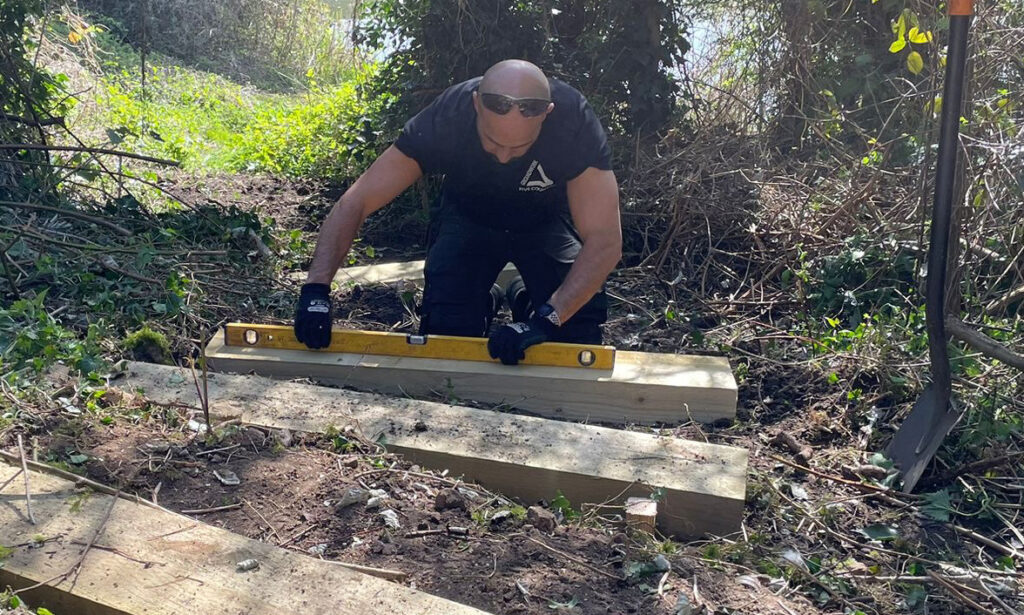 Man with sunglasses on forehead and wearing black, hunched over a measuring stick with wooden planks in outdoor space.