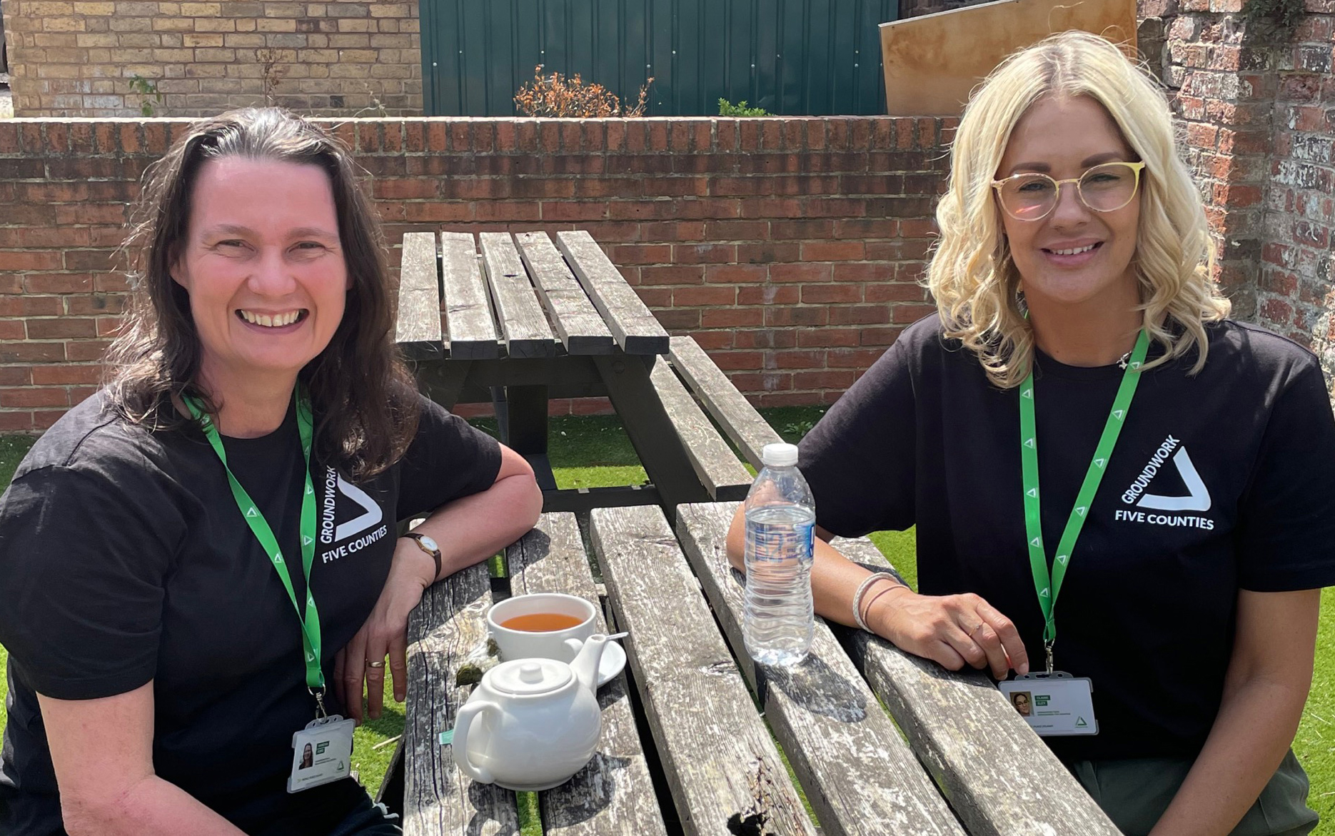 Two women, one brunette and one blonde, sitting on wooden bench with tea and water on the table smiling at the camera.