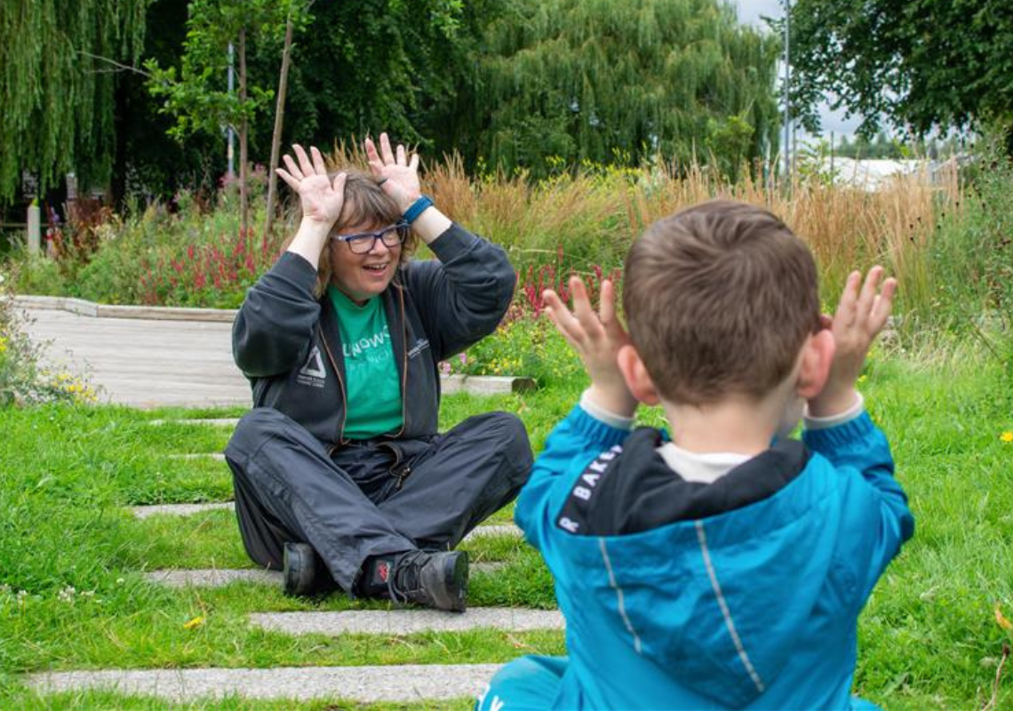 Young woman making funny faces at a small child, who has his back to the camera. They are both outdoors in some green space