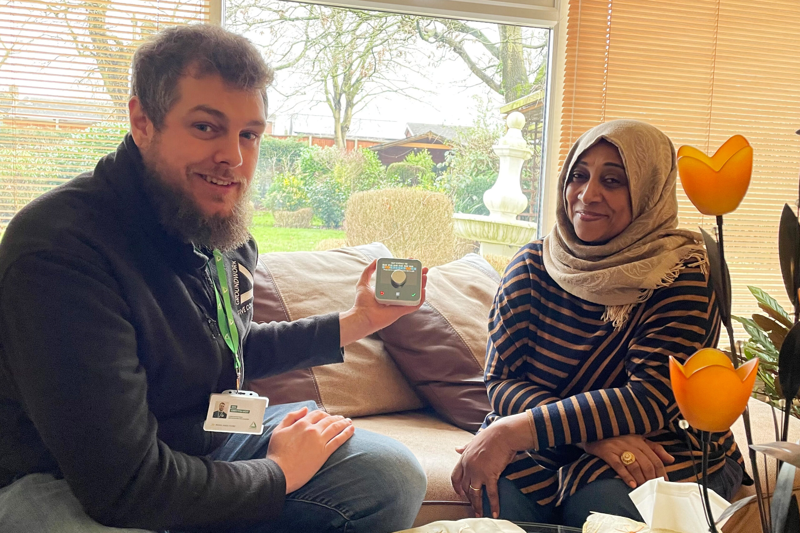 Man wearing black jacket and green lanyard sitting on sofa with woman in stripy top and grey hijab, holding smart thermostat and smiling at camera.