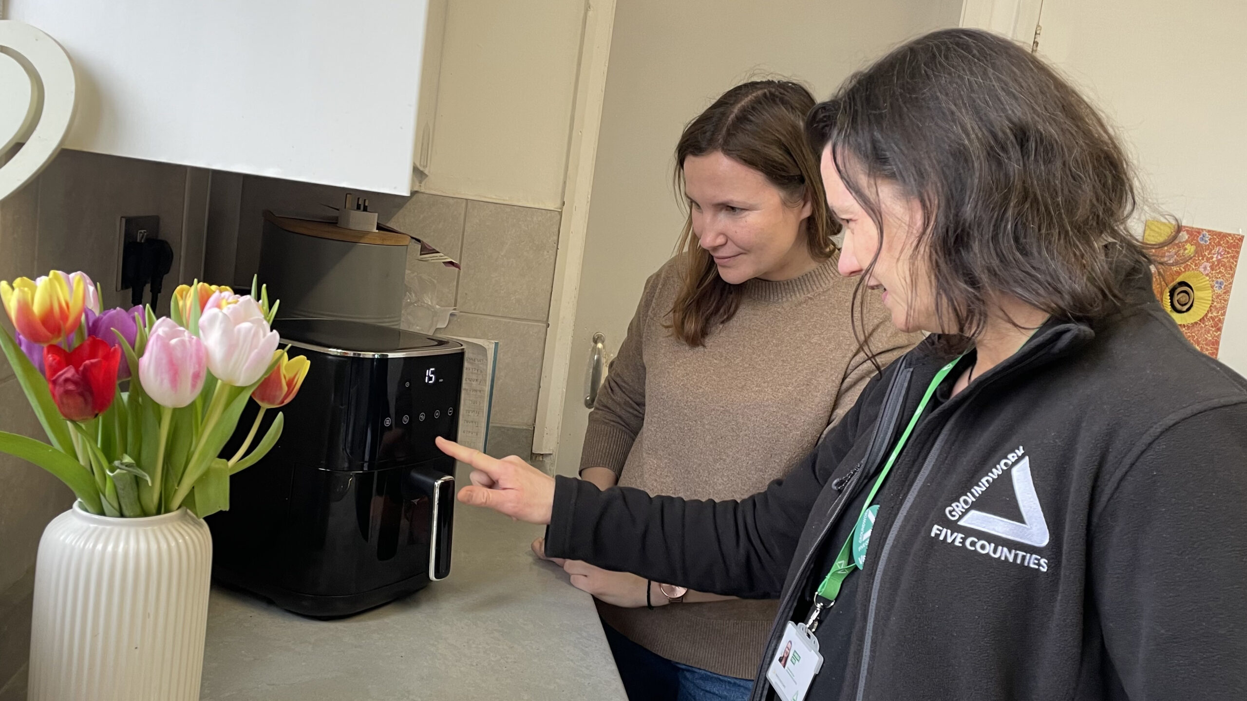 Two brunette women looking at air fryer on kitchen counter, with white vase and flowers in foreground.