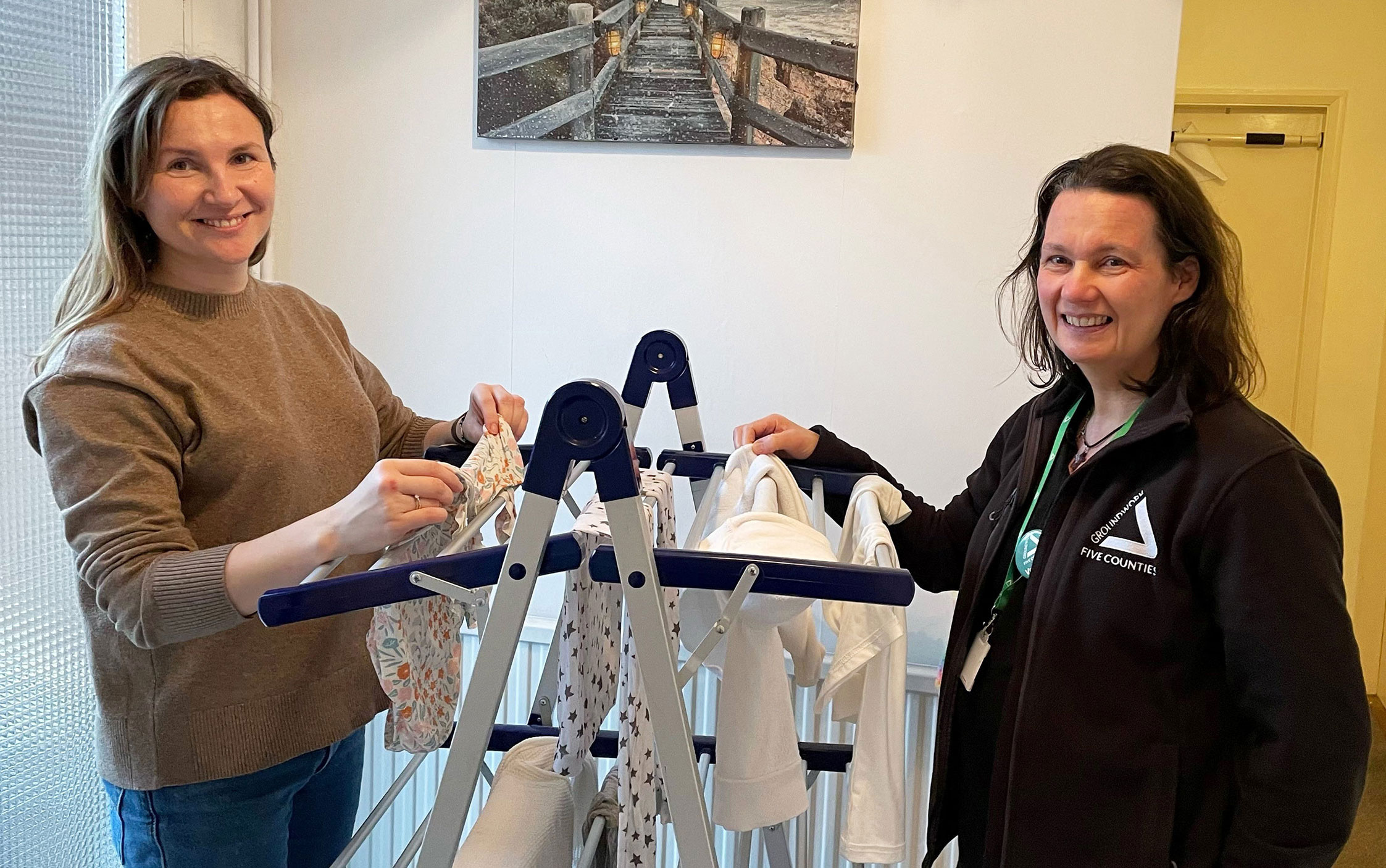 Two brunette women with clothes dryer and clothing items draped over, smiling at the camera.