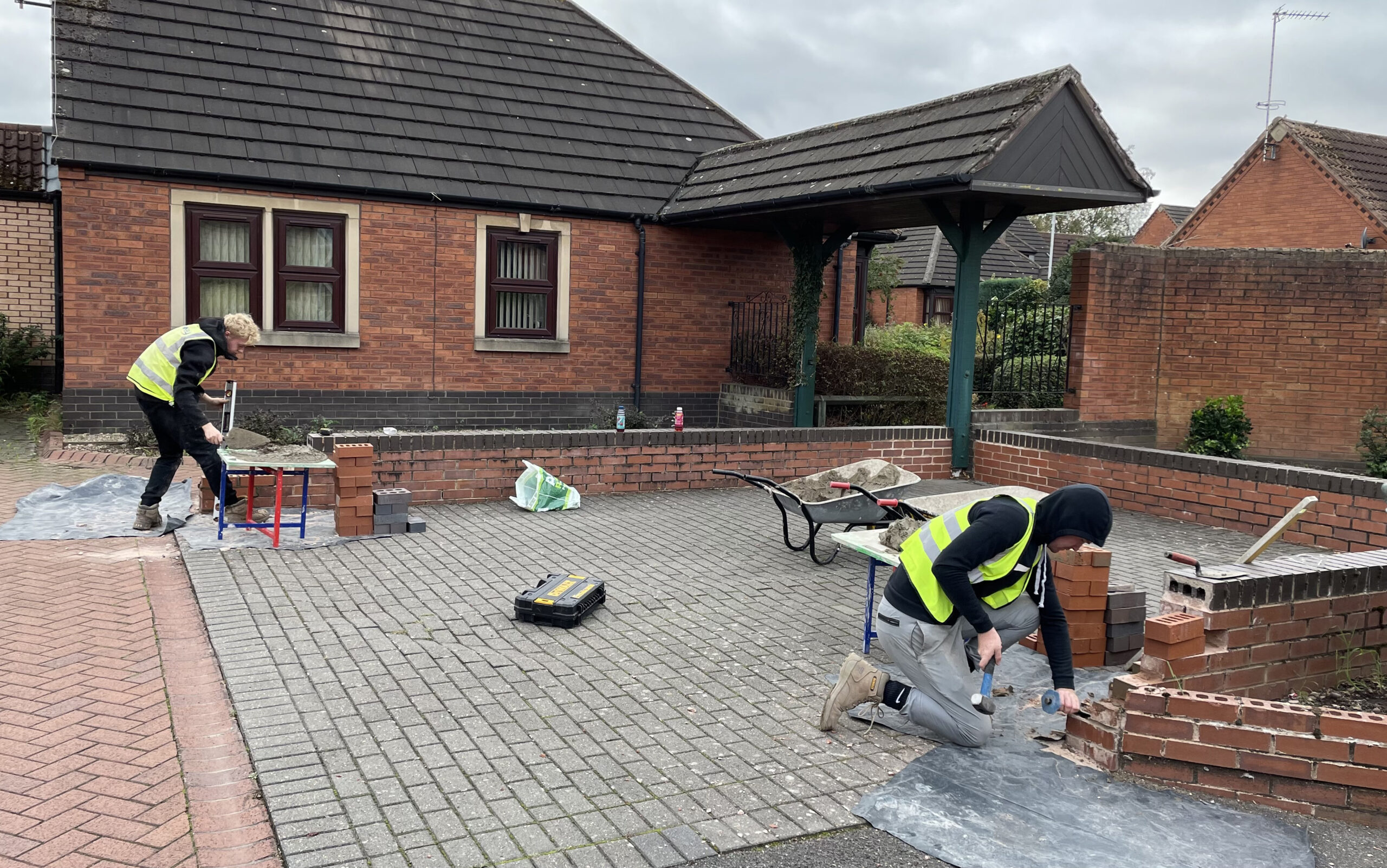Two young men wearing yellow high visibility vests doing brickwork on low walls in residential area.