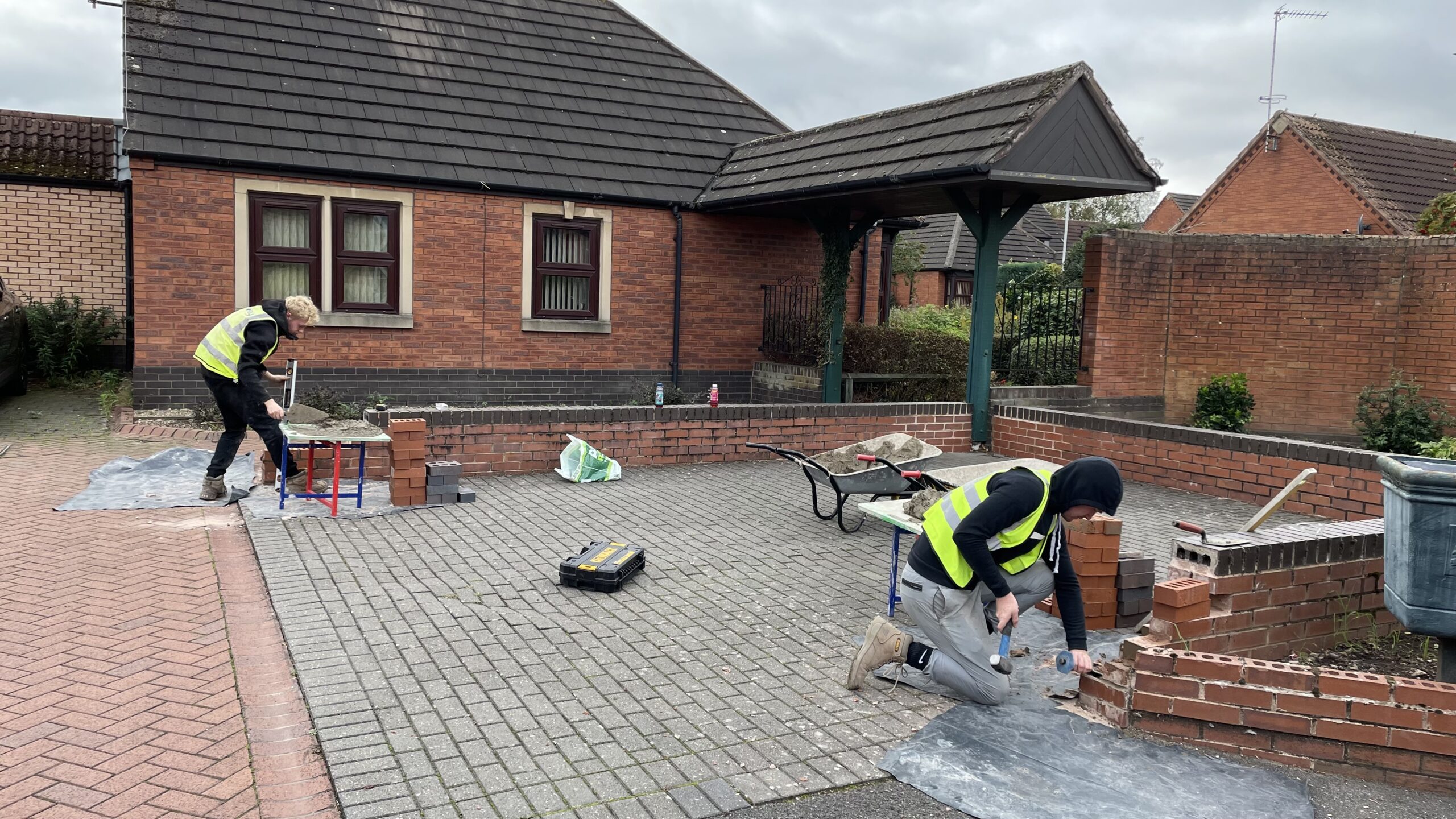 Two young men wearing yellow high visibility vests doing brickwork on low walls in residential area.