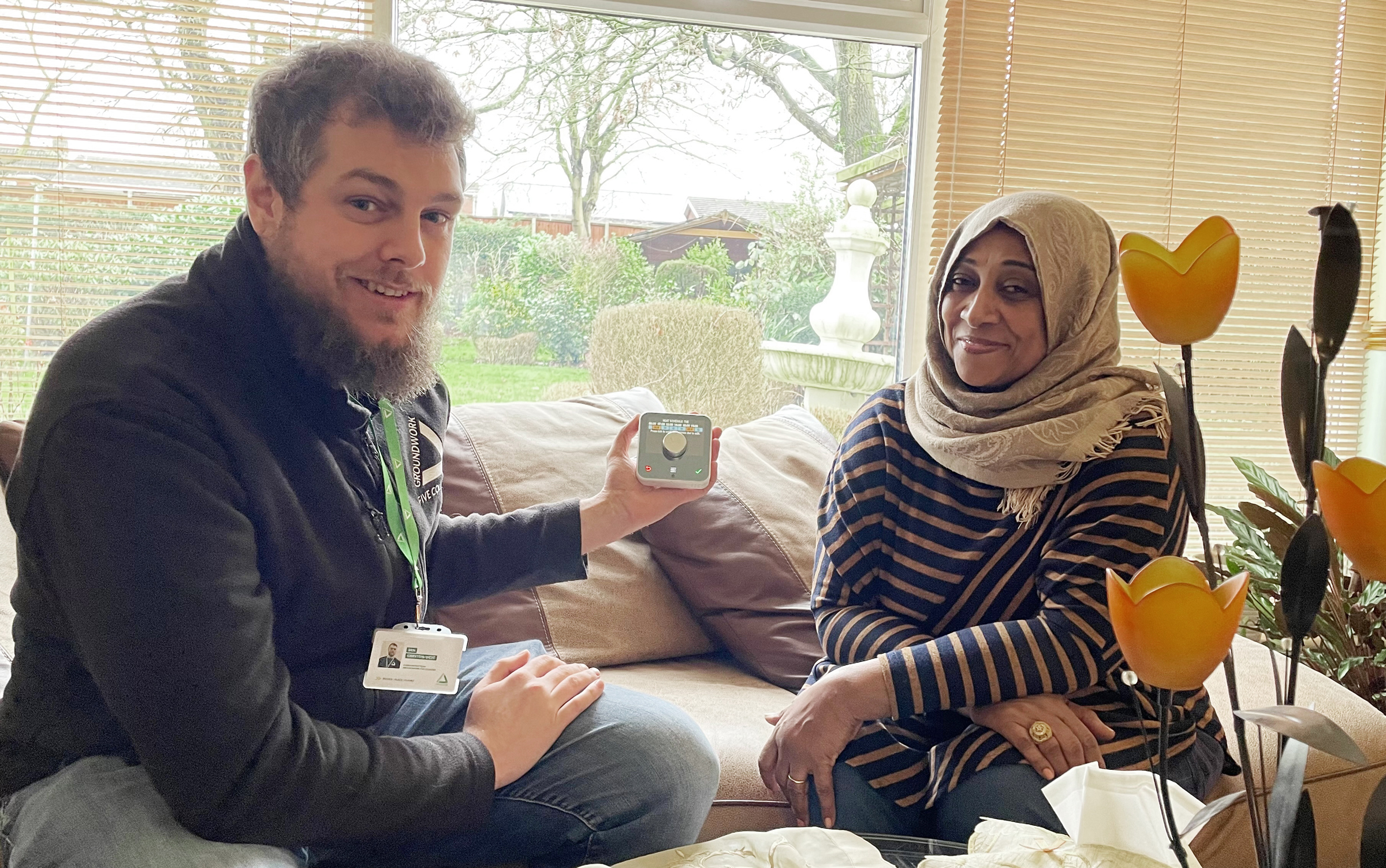 Man wearing black fleece and green lanyard sitting on sofa with woman in grey hijab and striped top. Man is holding a heating programmer, there are orange flowers in the foreground.