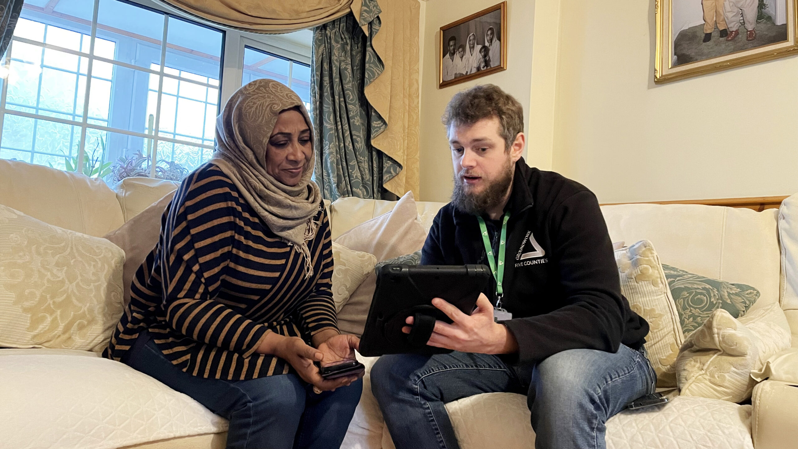 Woman wearing grey hijab and striped top sitting on cream sofa with man wearing black fleece and green lanyard, looking at tablet together,