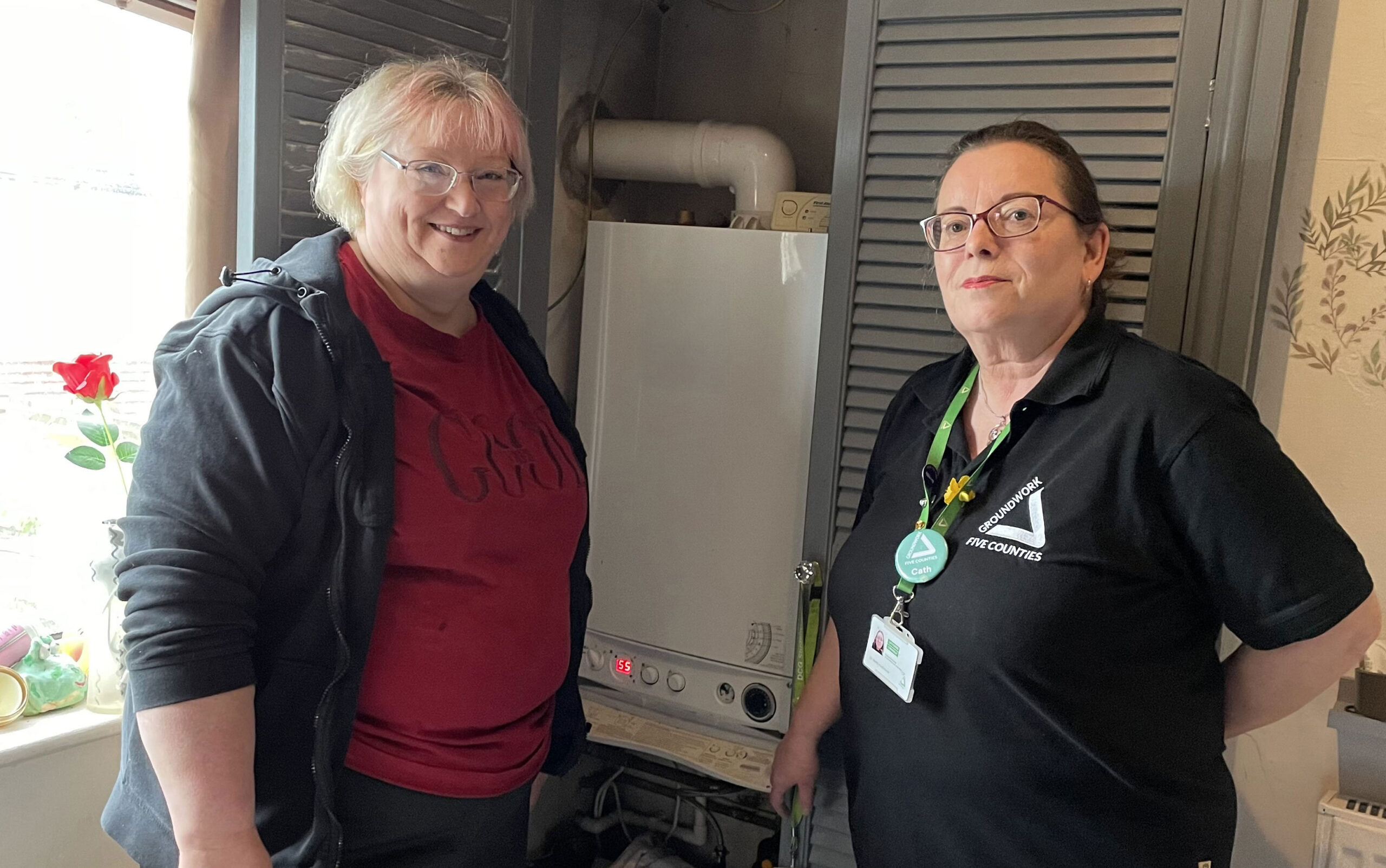 Two women standing on front of cupboard with boiler. On the left a blonde wearing a red top and black jacket, brunette on the right wearing a black top with white triangle logo and green lanyard.