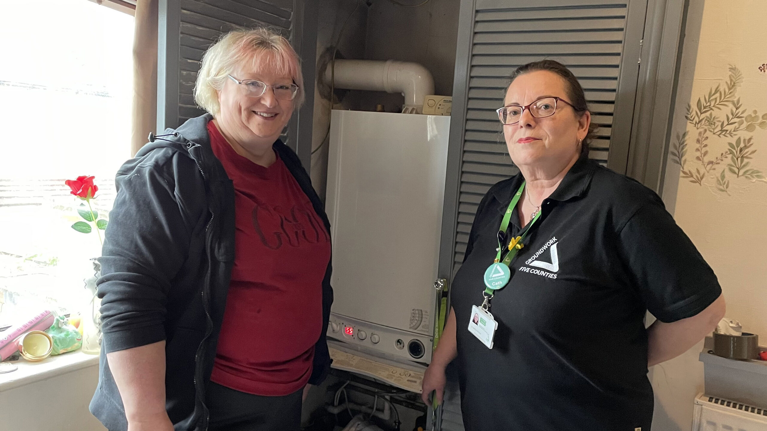 Two women standing on front of cupboard with boiler. On the left a blonde wearing a red top and black jacket, brunette on the right wearing a black top with white triangle logo and green lanyard.