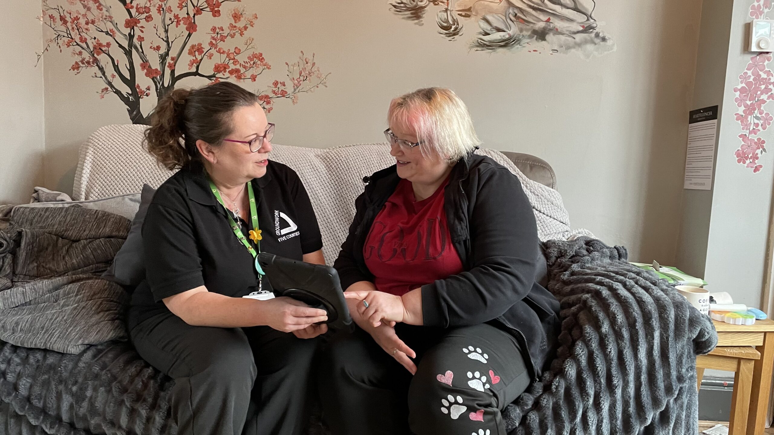 Two women sitting on grey sofa in cosy living room looking at tablet device.