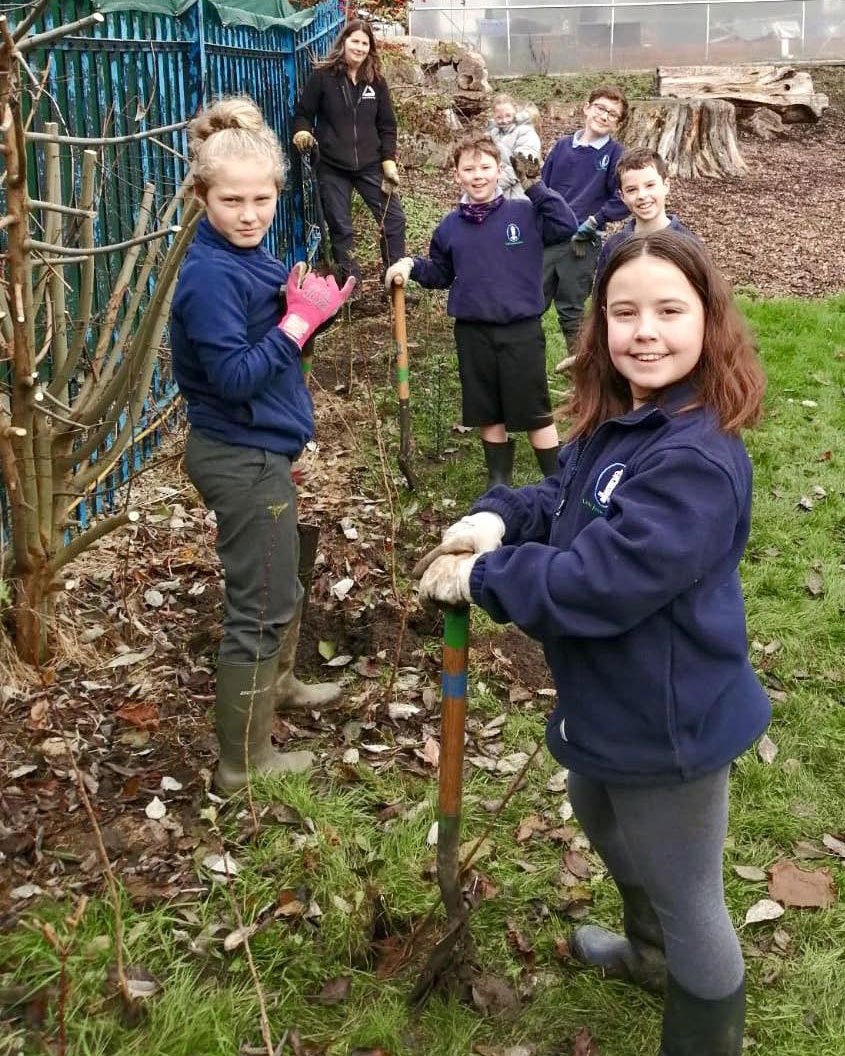 Group of young children with shovels in outdoor space with brunette woman in background