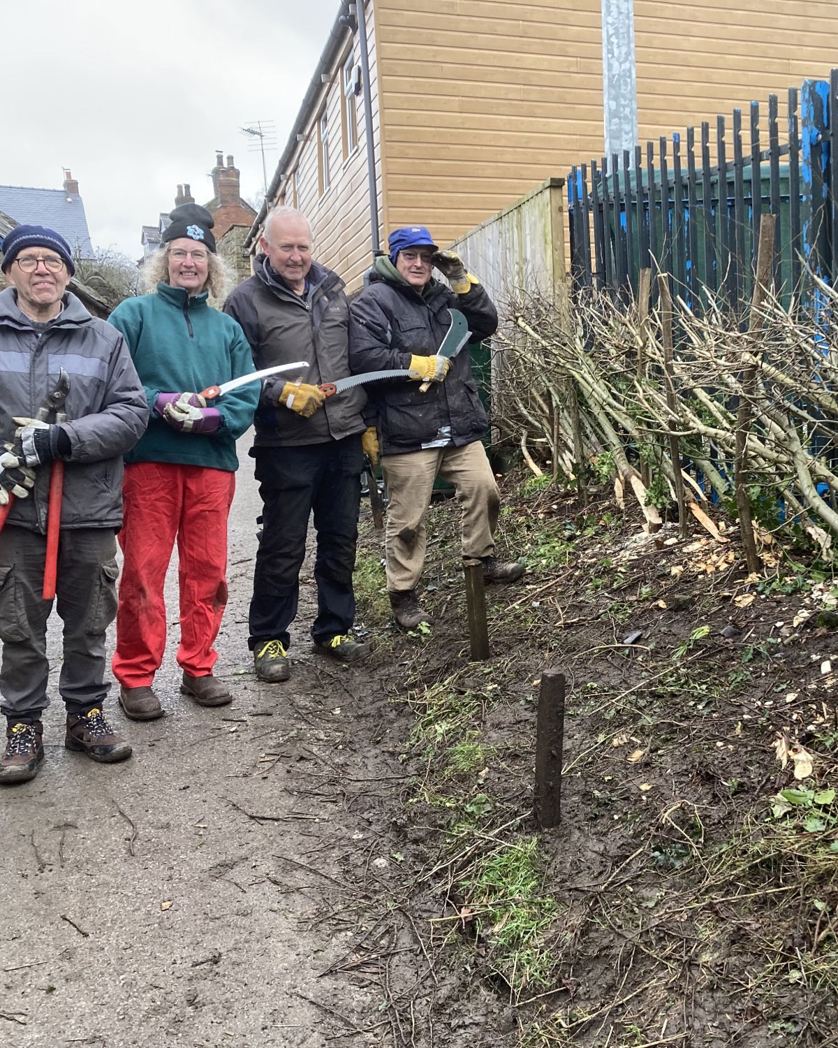 Three men and a woman holding gardening tools by hedge in residential area