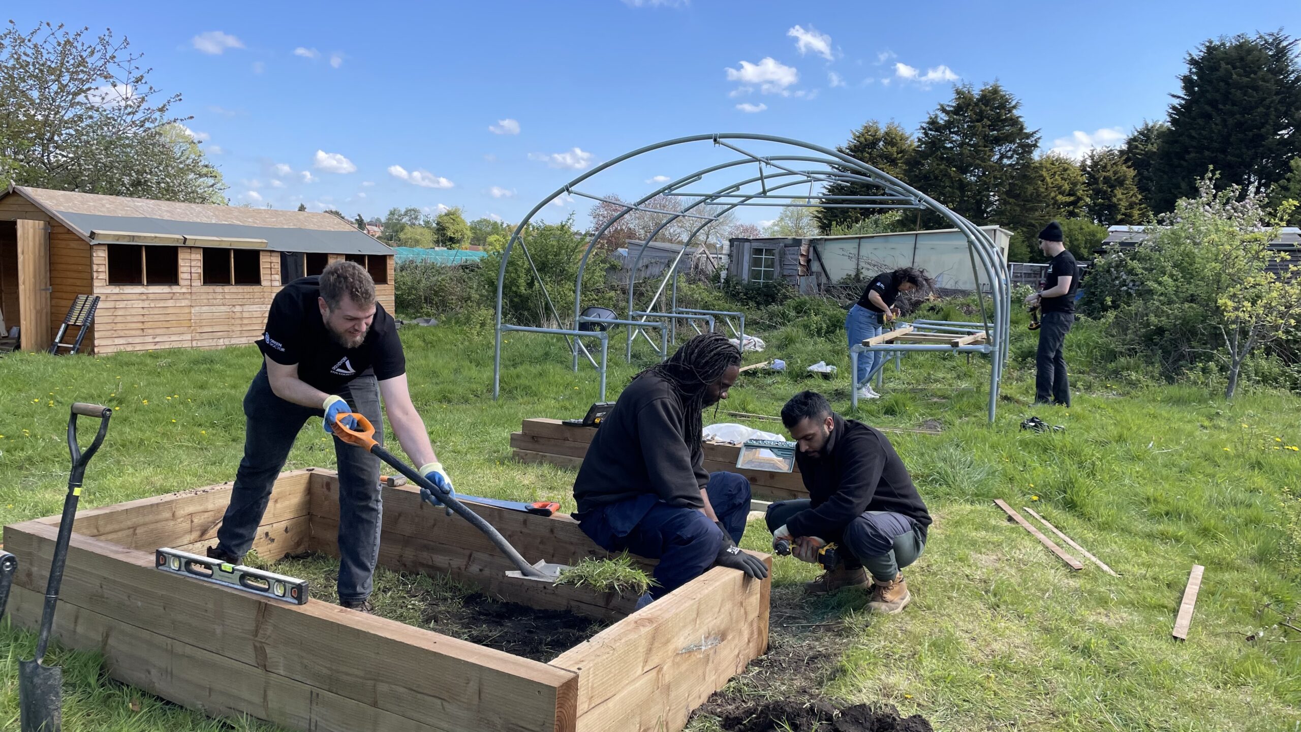 Three men working on wooden raised bed in allotment space, two people in the background working on polytunnel,
