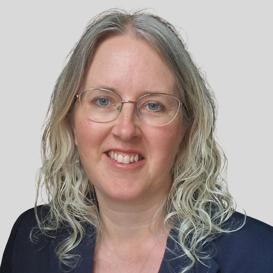 Woman with curly blonde-white hair, wearing glasses and dark blue jacket - on grey background.