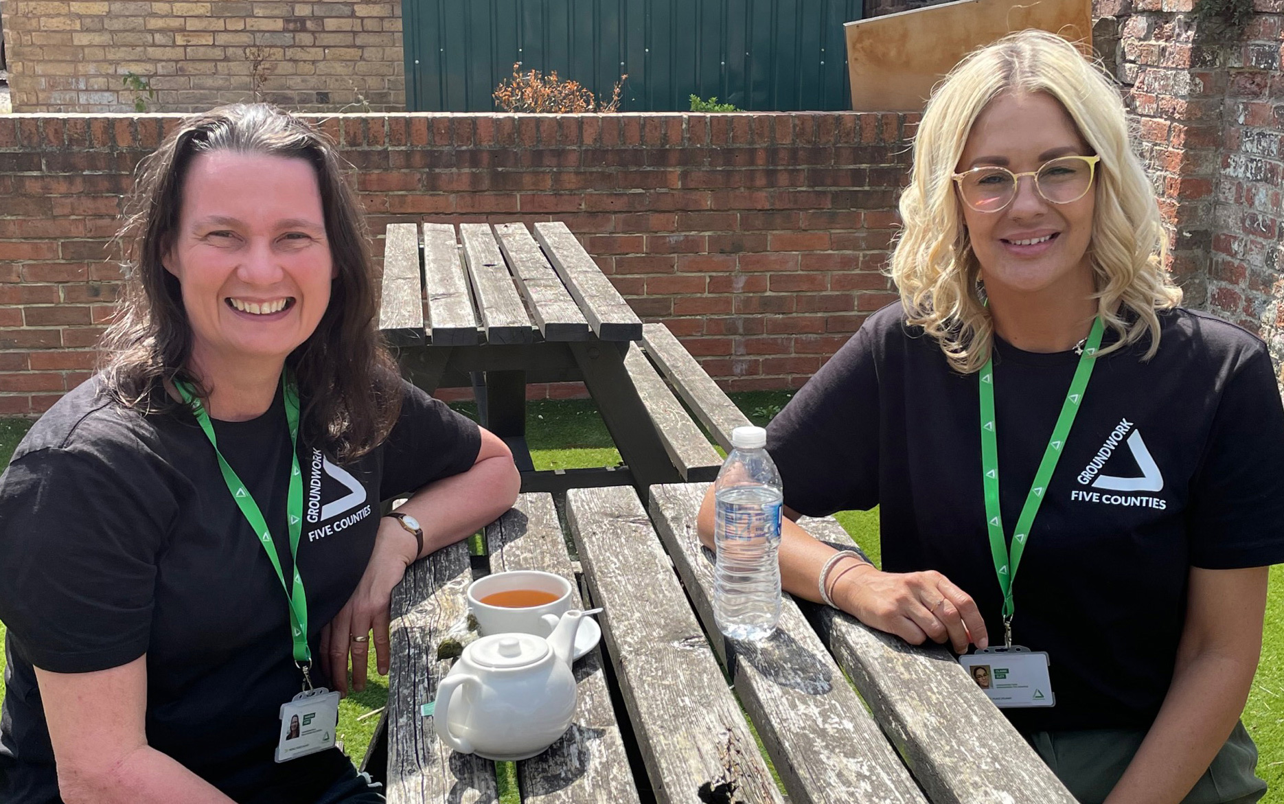 Two women, one brunette and one blonde, sitting on wooden bench with tea and water on the table smiling at the camera.