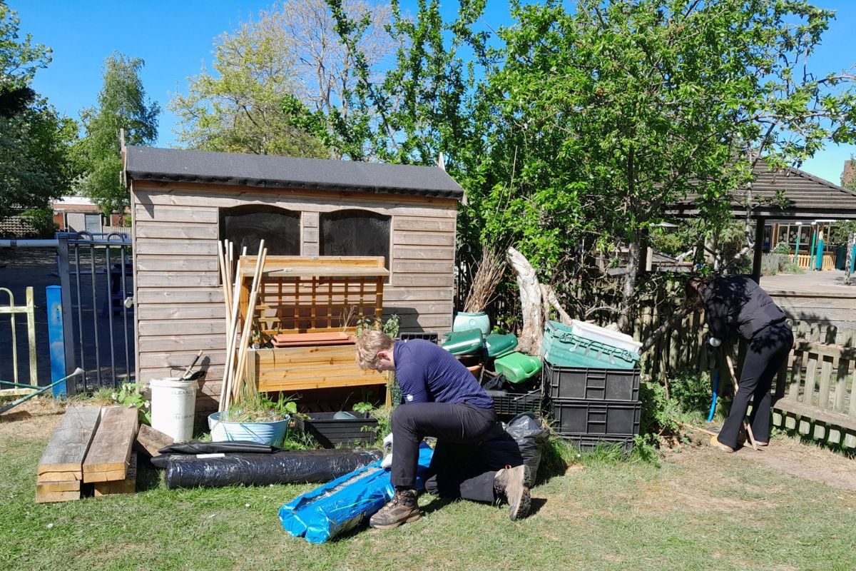 2 Volunteers helping improve the outdoor play area, one is knealing opening a packet on the ground, the other is using tools near a fence.