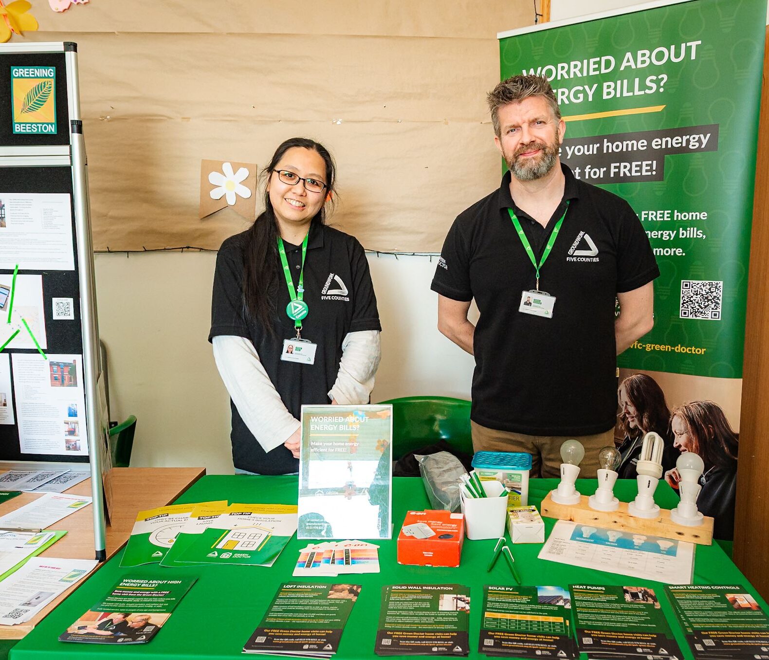 Woman and man wearing black T-shirts and green lanyards at community event with green banner, table cloth, and items on table.