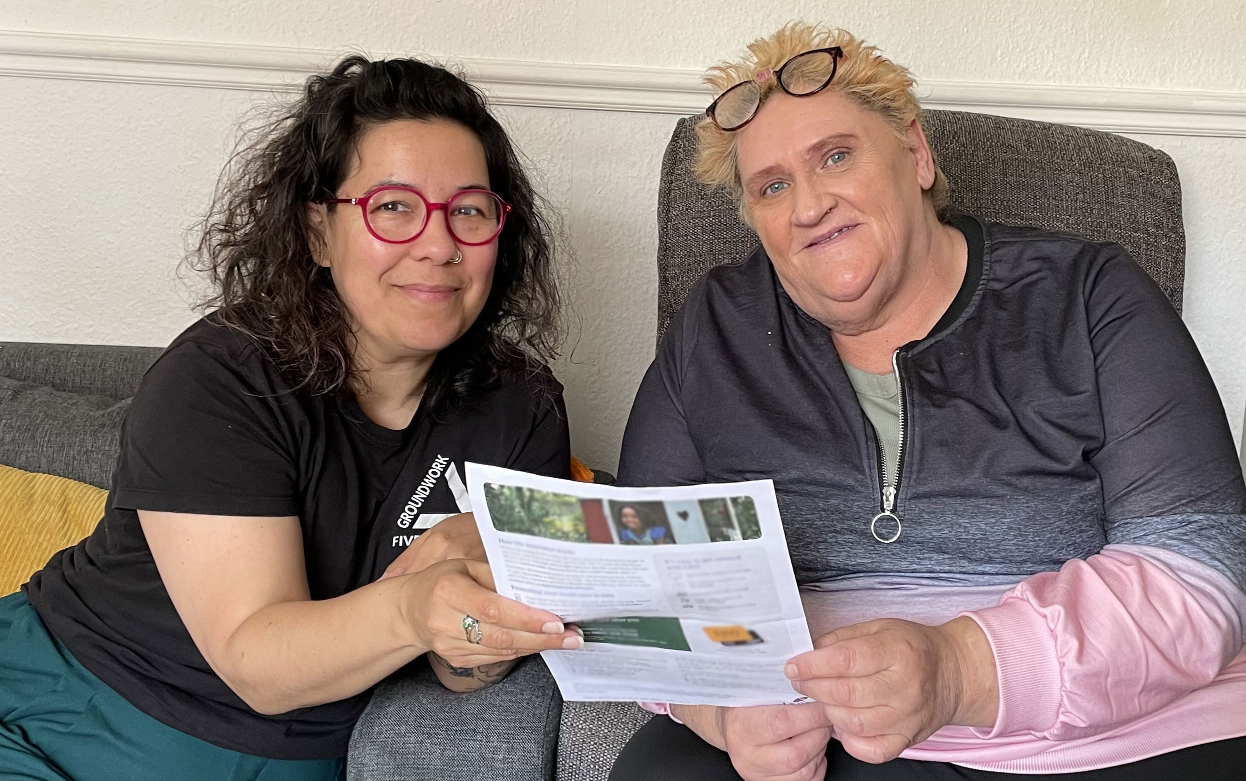 Two women holding piece of paper. Left woman has dark hair, pink glasses and is wearing a black top. Woman on the left has short spiky blonde hair, glasses on top of her head and a grey-pink top.