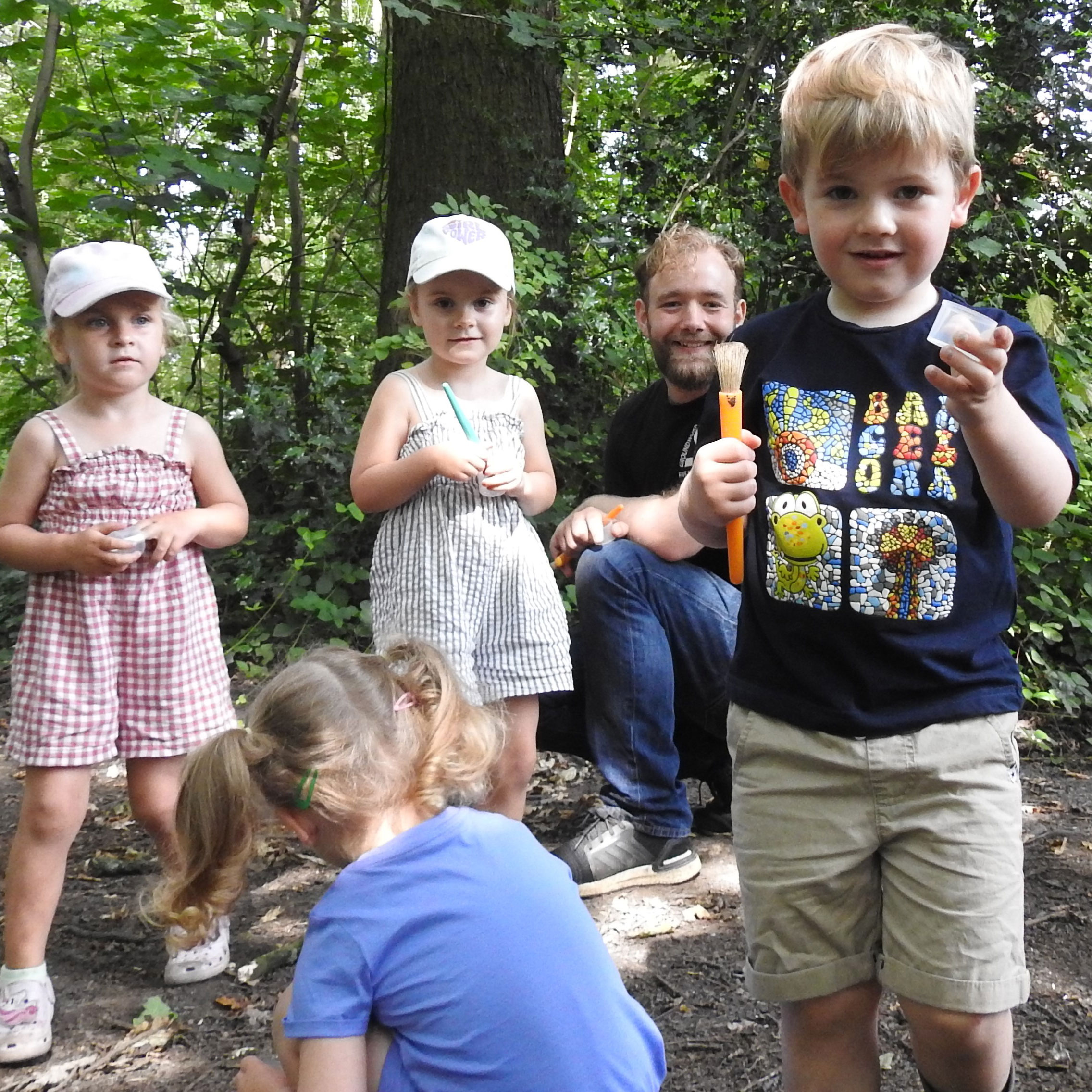 Group of young children and blonde man smiling at camera holding bug pots and brushes in outdoor space.