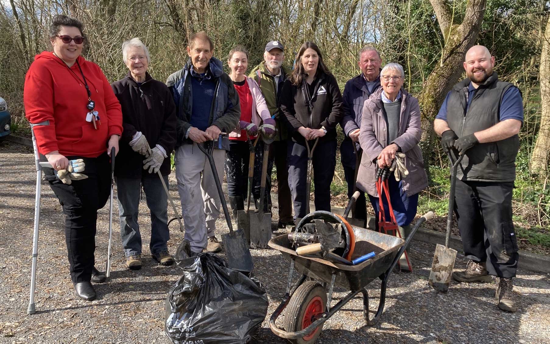 Group of people standing in front of woodland with wheelbarrow, tools, and black binbags.
