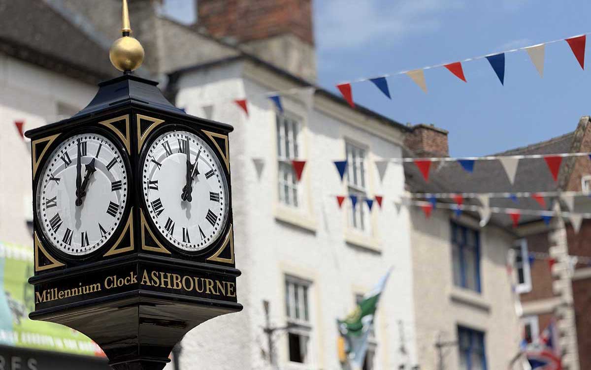 Black clock with white face and gold lettering 'Millennium Clock Ashbourne' in foreground with blurred background of street with red white and blue bunting.