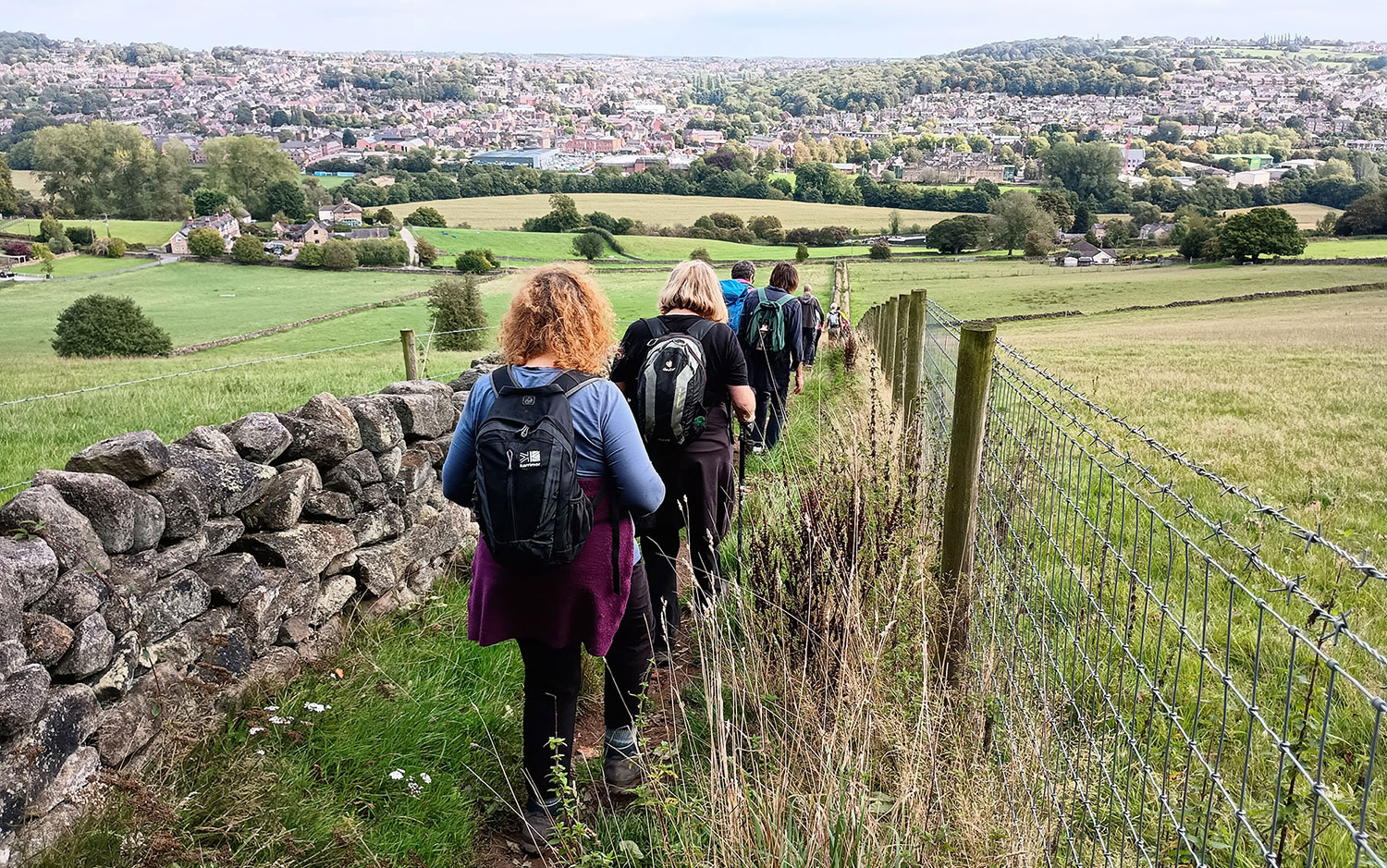 Group of people and walking down path by fencing and stone wall, with rolling hills and town in background