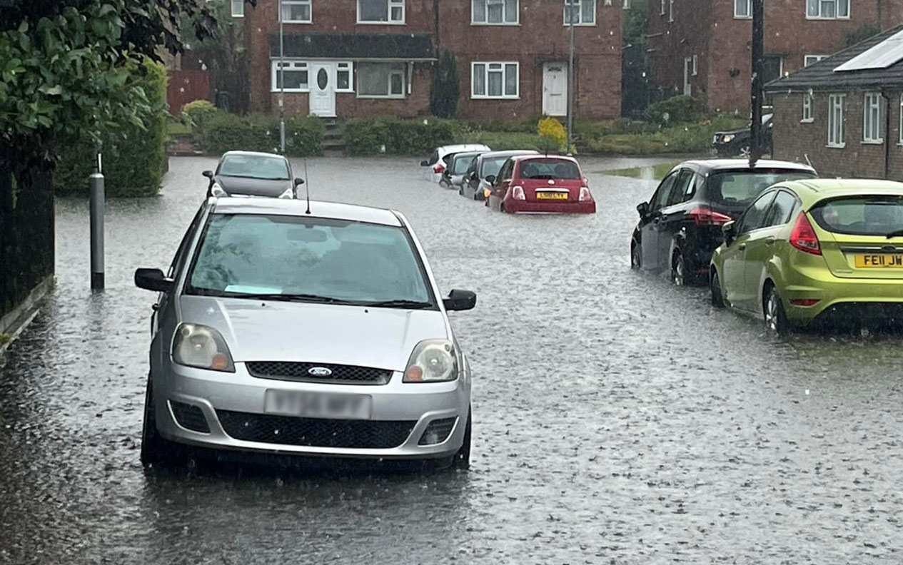 Flooded street with cars parked either side, the water is over the bumpers in some places