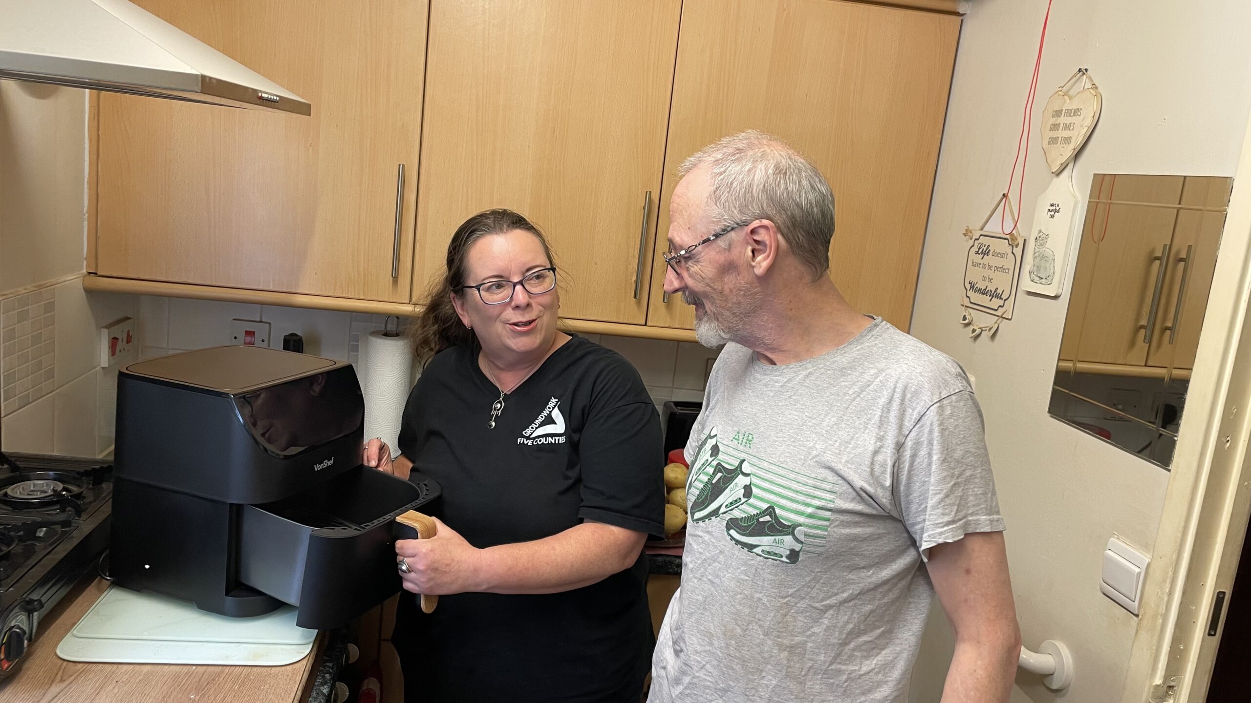 Brunette woman wearing black T-shirt and man wearing grey T-shirt standing in kitchen looking at air fryer