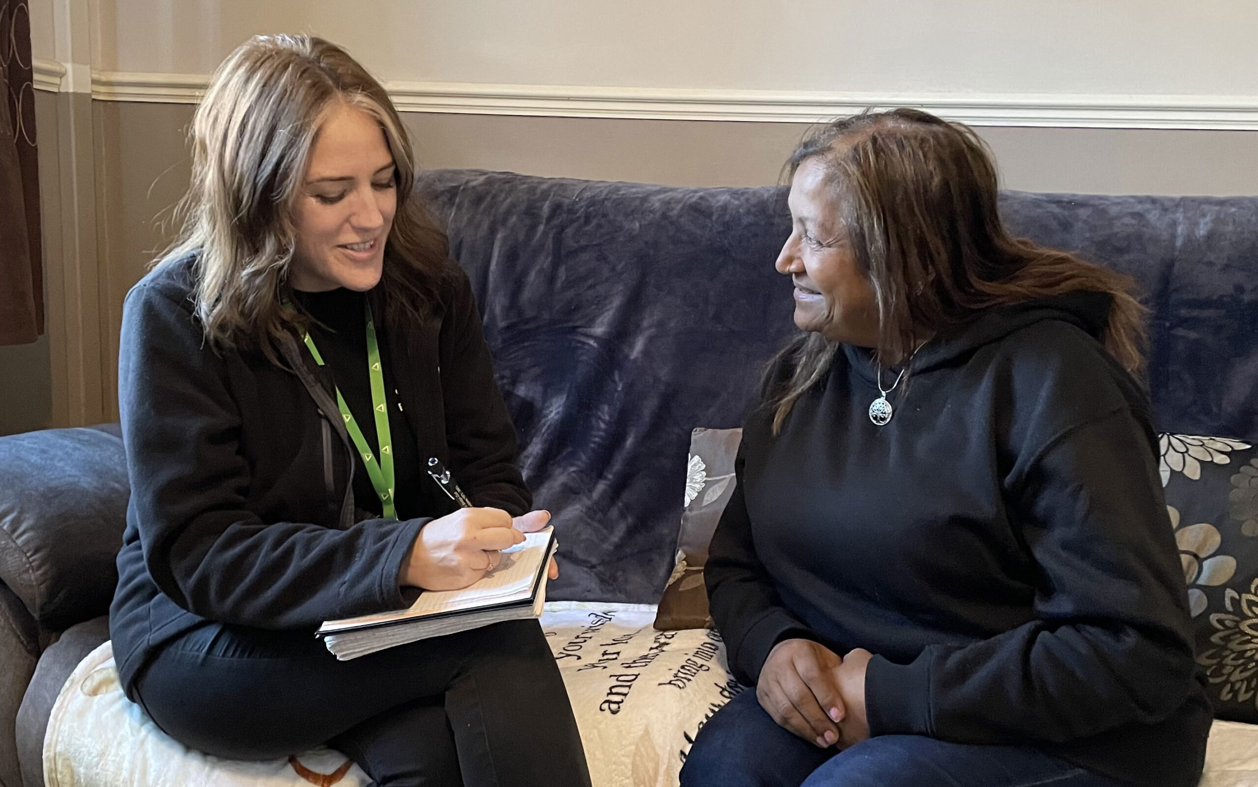 Two women sitting on sofa, talking and smiling. Woman on the left is taking notes on a notepad.