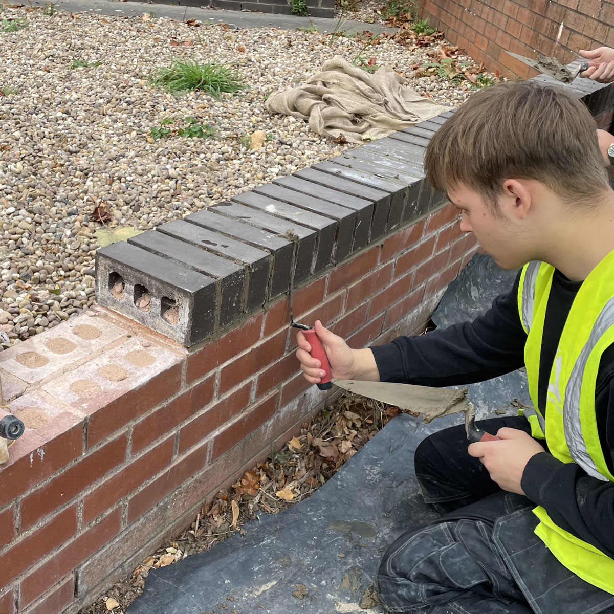 Young man wearing yellow high visibility vest and black top working on brick wall in outdoor environment