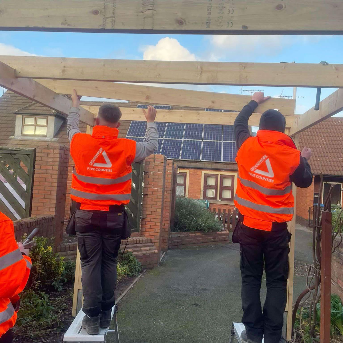 Two people wearing orange high visibility jackets placing wooden beam in outdoor environment