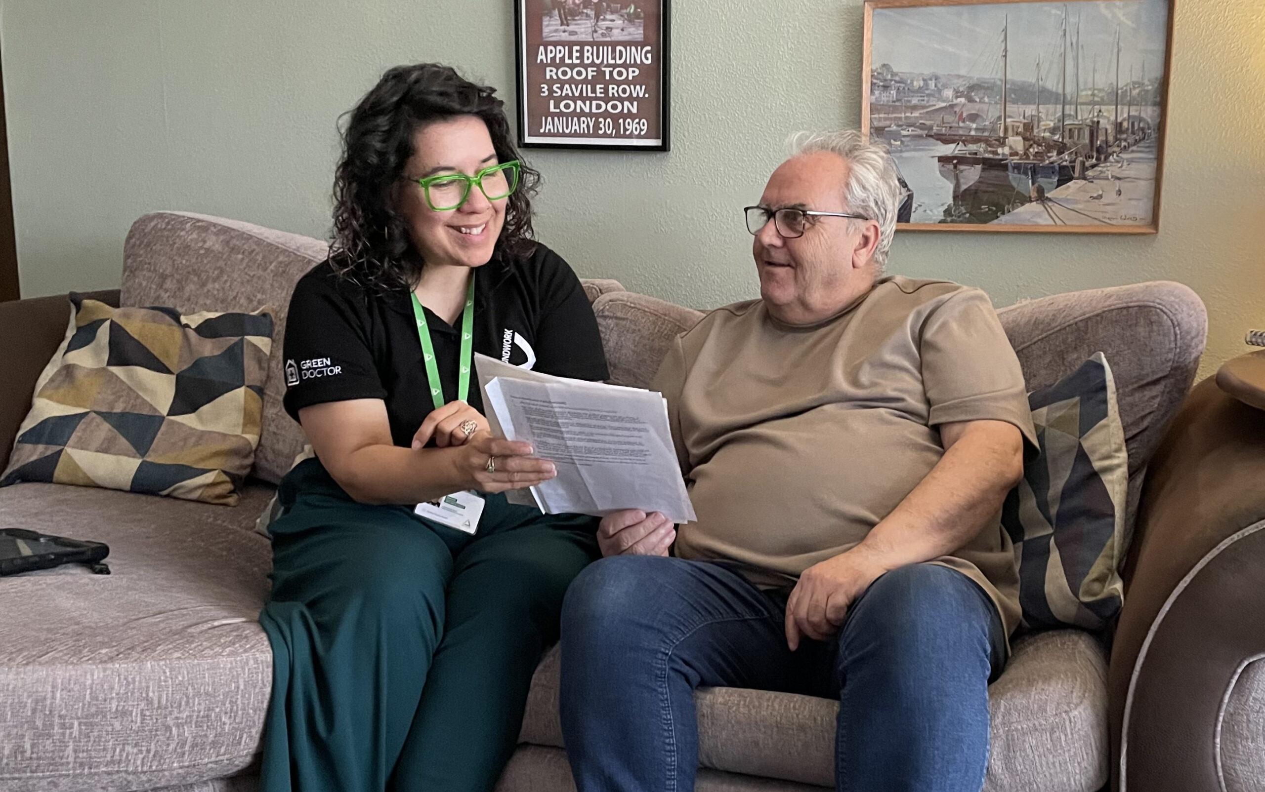 Woman wearing black top and green lanyard sitting on sofa with man wearing glasses and beige top, looking at paperwork together.