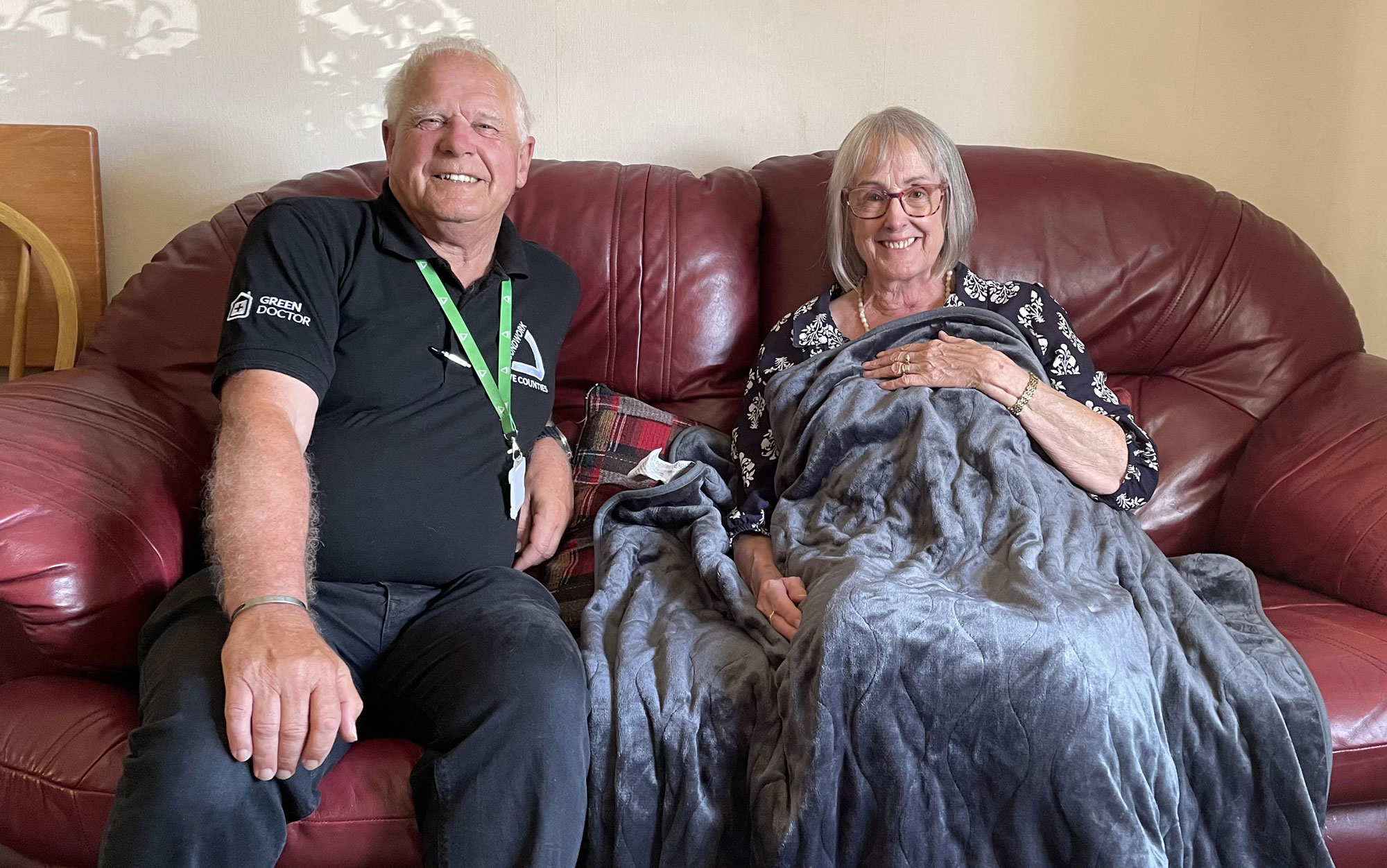 Man and woman sitting on dark red leather sofa, the woman holds a grey blanket over herself.