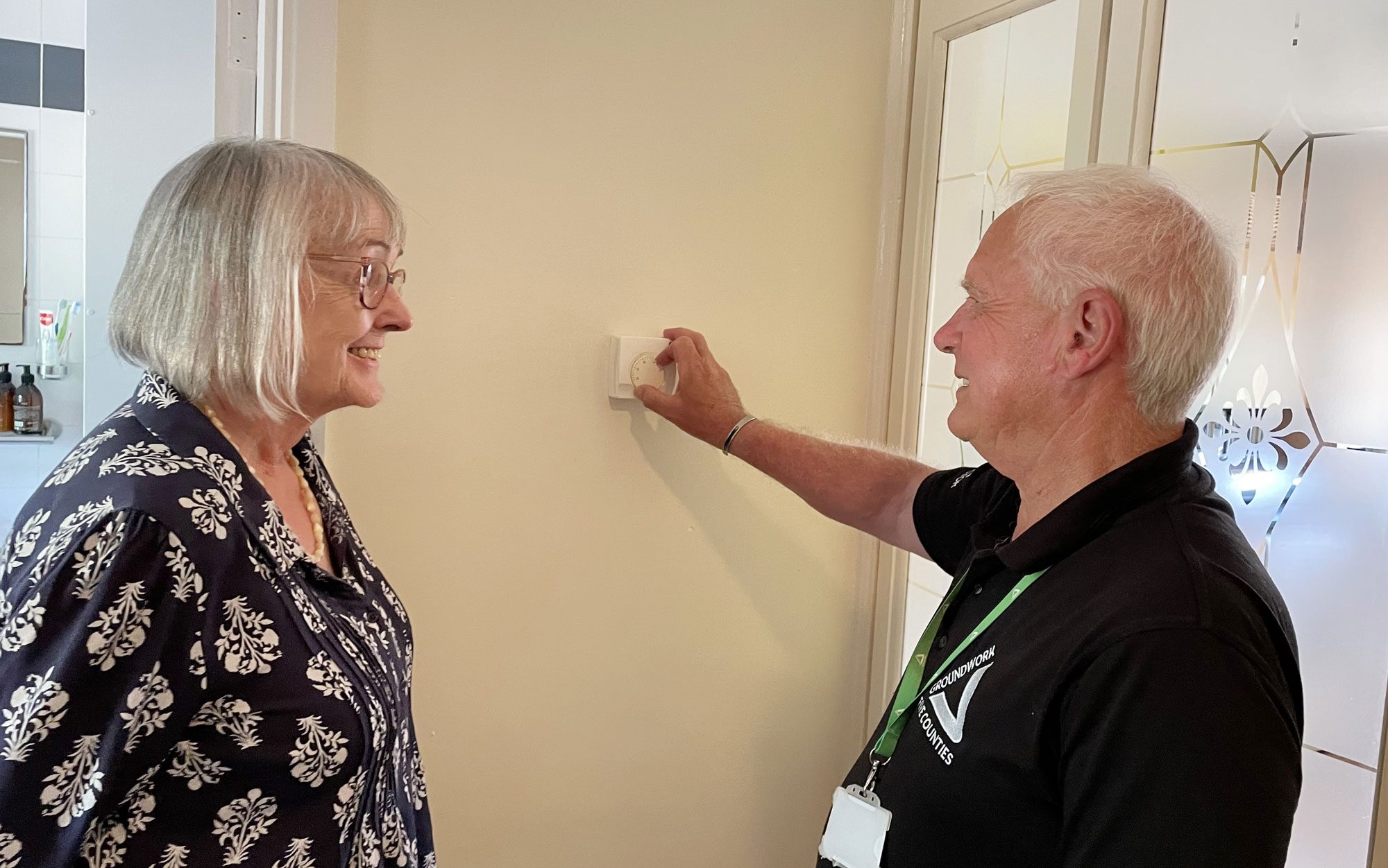 Woman and man standing by beige wall, man is adjusting a heating thermostat mounted on wall.